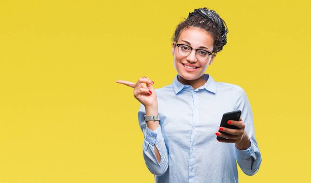 Jeune fille d'affaires afro-américaine cheveux tressés en utilisant un smartphone sur fond isolé très heureux pointant avec la main et le doigt sur le côté
 - Photo, image