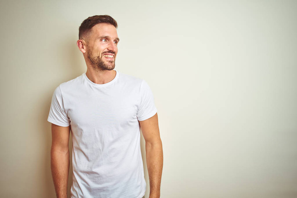 Young handsome man wearing casual white t-shirt over isolated background looking away to side with smile on face, natural expression. Laughing confident. - Foto, immagini