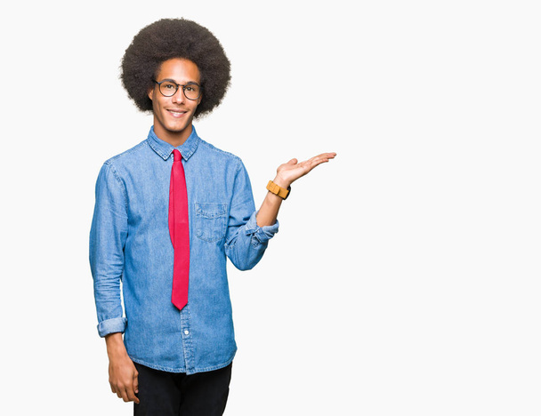 Young african american business man with afro hair wearing glasses and red tie smiling cheerful presenting and pointing with palm of hand looking at the camera. - Foto, afbeelding