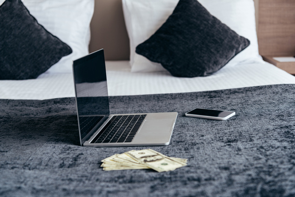 laptop with blank screen, smartphone and dollar banknotes on bed in hotel room - Фото, зображення