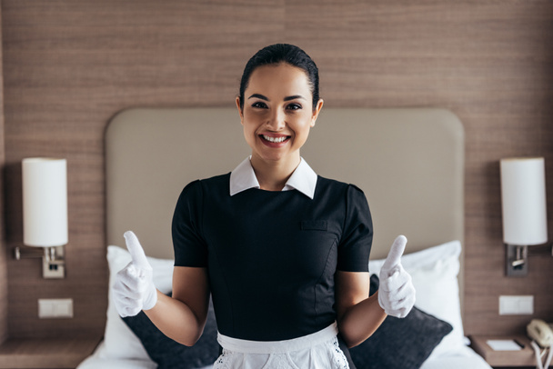 front view of pretty smiling maid in white gloves and apron looking at camera and showing thumbs up in hotel room - Fotografie, Obrázek