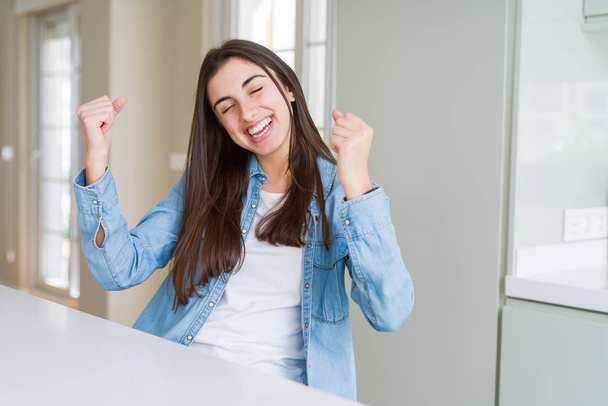 Belle jeune femme assise sur une table blanche à la maison très heureuse et excitée faisant geste gagnant avec les bras levés, souriant et criant pour le succès. Concept de célébration
. - Photo, image