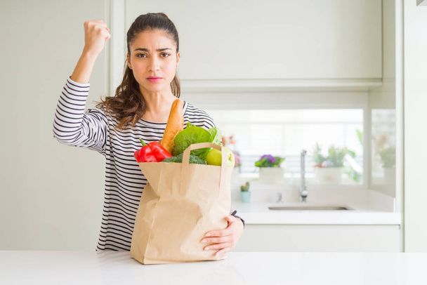 Young woman holding paper bag full of groceries annoyed and frustrated shouting with anger, crazy and yelling with raised hand, anger concept - Foto, afbeelding