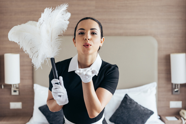 front view of smiling maid in white gloves holding duster and sending air kiss near bed in hotel room - Foto, Imagem