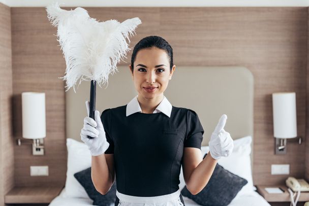 Vorderansicht des lächelnden Dienstmädchens in weißen Handschuhen, das Staubwedel hält und den Daumen in der Nähe des Schlafes im Hotelzimmer zeigt - Foto, Bild