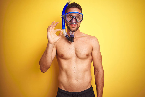 Young handsome shirtless man wearing diving goggles over isolated yellow background doing ok sign with fingers, excellent symbol - Photo, image