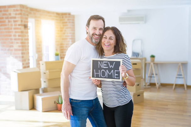 Seniorenpaar mittleren Alters, das in ein neues Haus zieht, glücklich verliebt lächelnd mit einer Tafel mit neuem Heimtext in der Hand - Foto, Bild