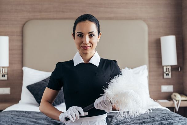 front view of smiling maid in white gloves holding duster near bed and looking at camera - Zdjęcie, obraz