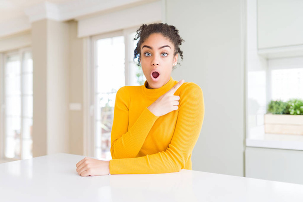 Mulher americana africana bonita com cabelo afro vestindo uma camisola amarela casual Surpreendido apontando com o dedo para o lado, boca aberta expressão surpreendente
. - Foto, Imagem