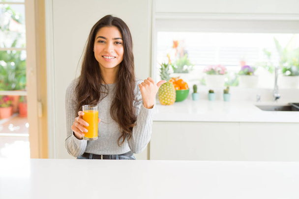 Giovane donna che beve un bicchiere di succo d'arancia fresco indicando e mostrando con il pollice verso l'alto sul lato con il viso felice sorridente
 - Foto, immagini