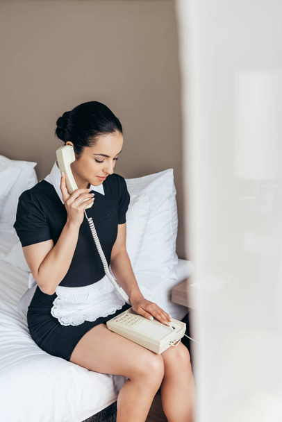 graziosa cameriera in grembiule bianco seduta sul letto e che parla al telefono in camera d'albergo
 - Foto, immagini