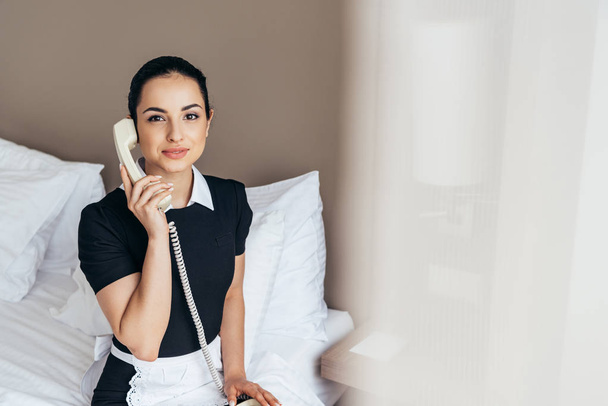 pretty maid in white apron sitting on bed and talking on telephone in hotel room - Fotoğraf, Görsel