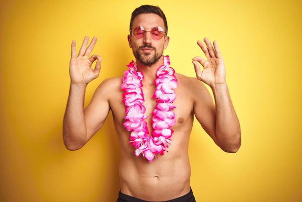 Young handsome shirtless man wearing sunglasses and pink hawaiian lei over yellow background relax and smiling with eyes closed doing meditation gesture with fingers. Yoga concept. - Photo, image