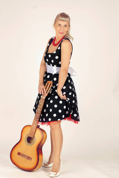 Mooie blonde vrouw in Pinup stijl, gekleed in een polka dot jurk, staat en houdt een akoestische gitaar voor haar, witte achtergrond - Foto, afbeelding