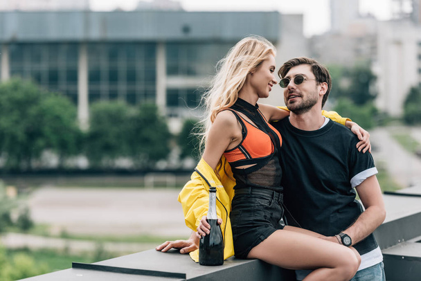 blonde woman with bottle and handsome man in glasses hugging and talking on roof  - Фото, изображение