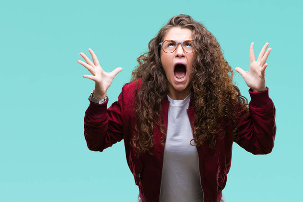 Beautiful brunette curly hair young girl wearing jacket and glasses over isolated background celebrating mad and crazy for success with arms raised and closed eyes screaming excited. Winner concept - Foto, imagen