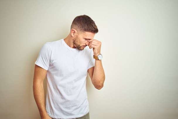 Jonge knappe man dragen casual wit t-shirt over geïsoleerde achtergrond moe wrijven neus en ogen, vermoeidheid en hoofdpijn gevoel. Stress en frustratie concept. - Foto, afbeelding