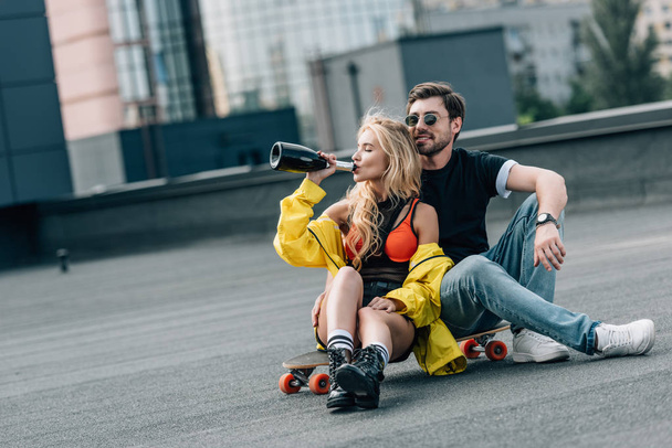 attractive woman drinking champagne and handsome man in glasses sitting on skateboard  - Fotoğraf, Görsel