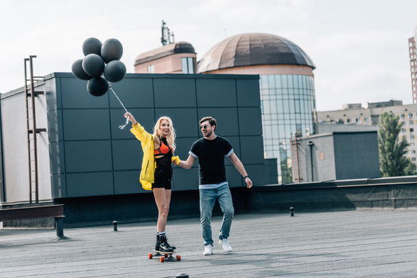 attractive woman with balloons skateboarding and man in glasses holding her hand - Фото, изображение