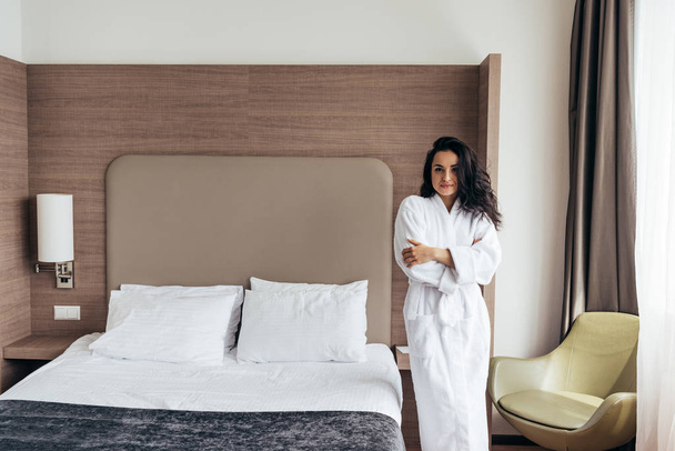 attractive brunette young woman in white bathrobe standing near bed in cozy bedroom - Foto, imagen
