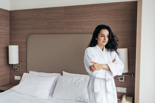attractive brunette young woman in white bathrobe standing near bed in cozy bedroom - Foto, Imagen