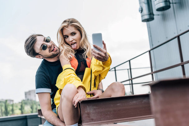 attractive and blonde woman sticking out tongue and taking selfie with handsome man     - Фото, изображение