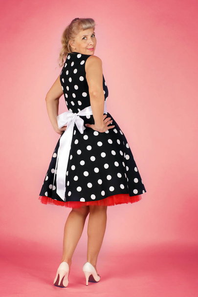 Beautiful blonde woman in a polka-dot dress flirty over her shoulder, pin-up style on a pink background - Foto, afbeelding