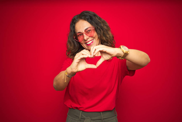 Middelbare leeftijd Senior vrouw het dragen van schattige hartvormige glazen over rode geïsoleerde achtergrond glimlachen in liefde tonen hart symbool en vorm met de handen. Romantisch concept. - Foto, afbeelding