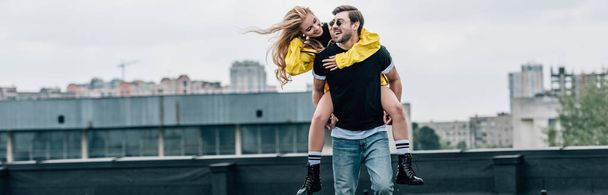 panoramic shot of attractive and blonde woman and handsome man hugging and playing on roof  - 写真・画像