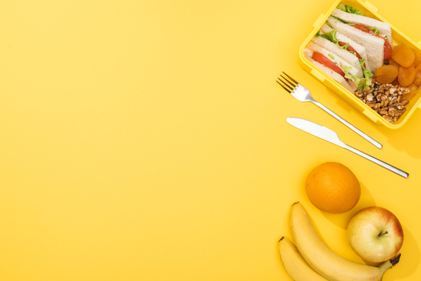 top view of lunch box with sandwiches, nuts, dried apricots, near fork, knife, oranges and bananas - Photo, Image