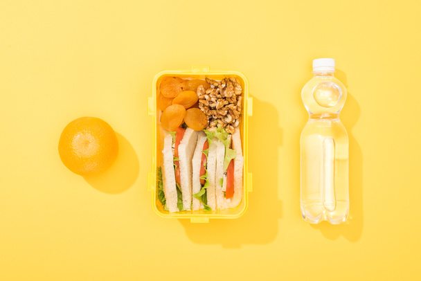 вид на обеденную коробку с бутербродами, орехами, сушеными абрикосами возле бутылки с водой и апельсином
 - Фото, изображение