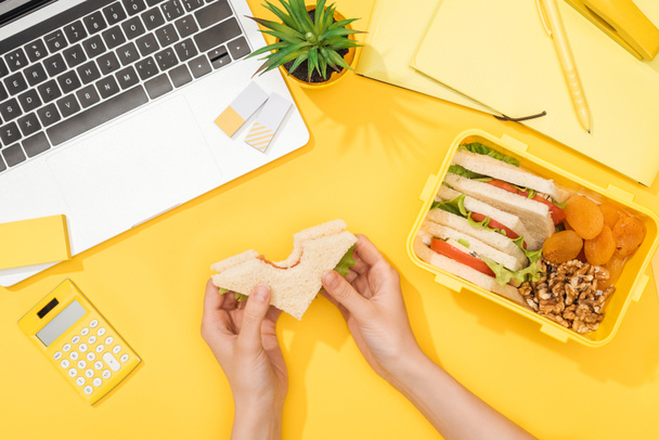 обрезанный вид женщины, держащей сэндвич в руках рядом с обеденной коробкой, ноутбуком и офисными принадлежностями
 - Фото, изображение