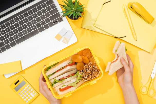 cropped view of woman holding sandwich in hand near lunch box, laptop and office supplies - Photo, Image