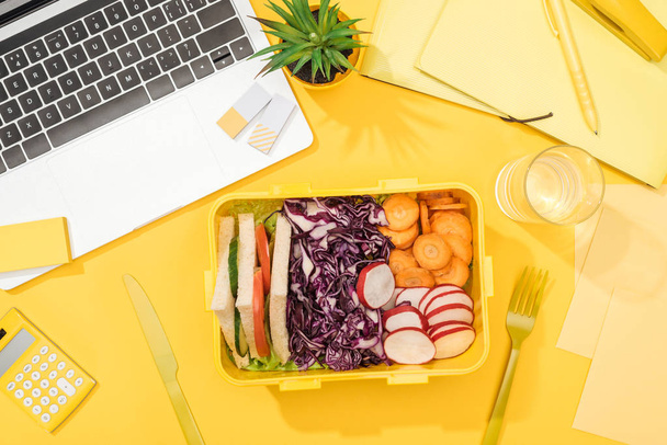 top view of lunch box with food near laptop, glass of water and office supplies - Photo, Image