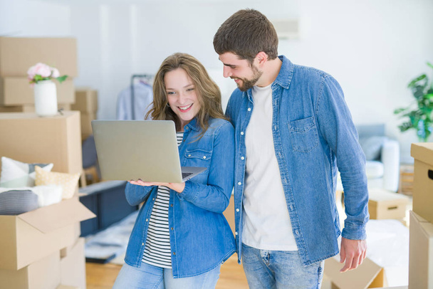 junges Paar mit Computer-Laptop steht in einem Raum um Kartons herum und freut sich über den Umzug in eine neue Wohnung - Foto, Bild