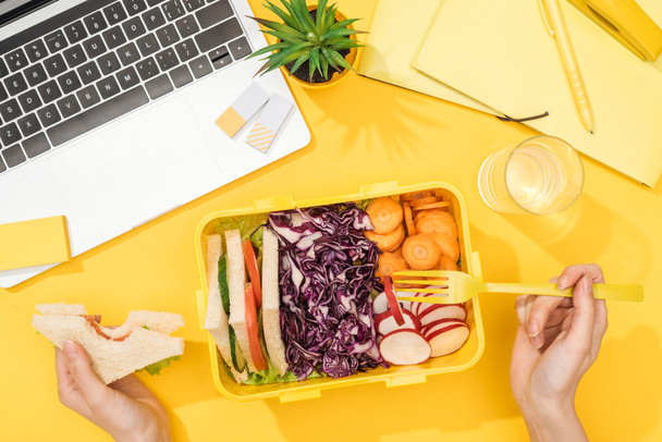 oříznutý pohled na ženu, která drží sendvič v ruce blízko polední krabice s jídlem, laptopem, skleněnými vodami a kancelářskou potřebami - Fotografie, Obrázek