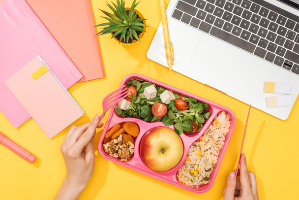 cropped view of woman holding fork over lunch box with food near laptop and office supplies - Photo, Image