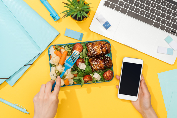 cropped view of woman holding smartphone near lunch box, laptop and office supplies - Photo, Image