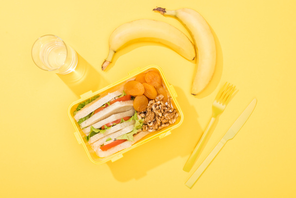 Draufsicht auf Lunchbox mit Lebensmitteln in der Nähe von Glas Wasser, Bananen und Plastikgeschirr - Foto, Bild