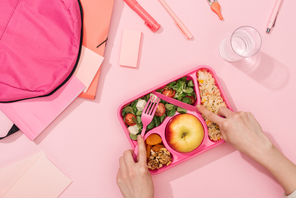 cropped view of woman hands with plastic utensils over lunch box with food near backpack and stationery - Photo, Image