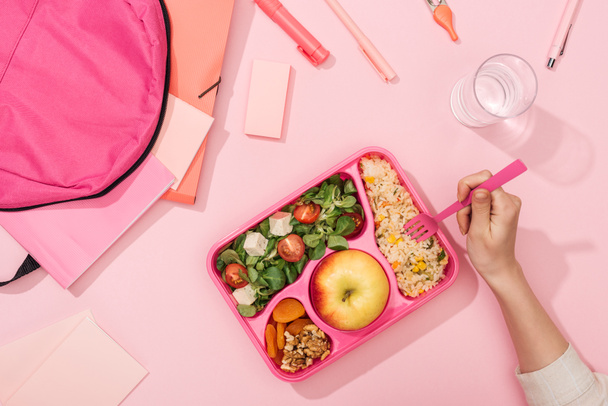 обрезанный вид женских рук с пластиковой посудой над обеденной коробкой с едой возле рюкзака и канцелярских принадлежностей
 - Фото, изображение