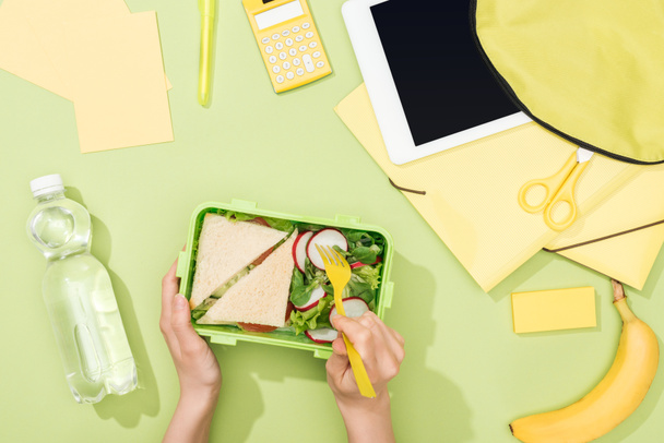 vista ritagliata di mani di donna con utensili di plastica sopra la scatola di pranzo con cibo vicino allo zaino, tablet digitale, bottiglia d'acqua e cancelleria
 - Foto, immagini