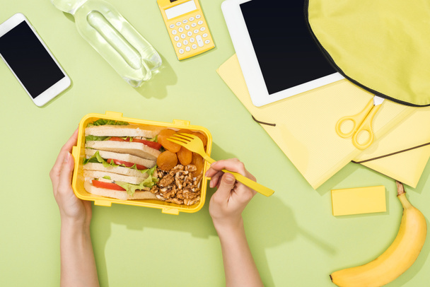 cropped view of woman hands with plastic utensils over lunch box with food near backpack, digital tablet, bottle of water and stationery - Photo, Image