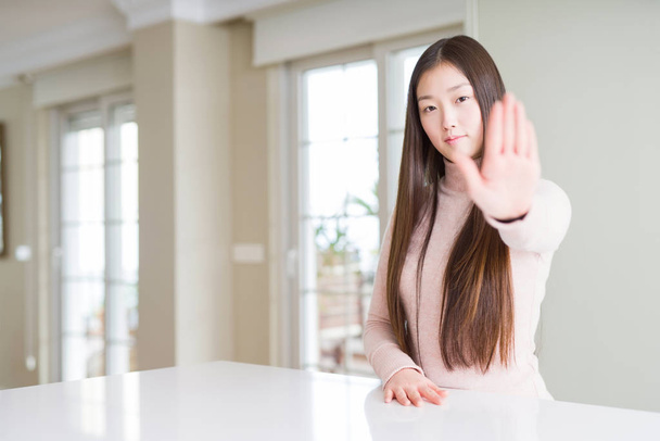Belle femme asiatique portant un pull décontracté sur une table blanche faisant arrêter chanter avec la paume de la main. Expression d'avertissement avec geste négatif et sérieux sur le visage
. - Photo, image