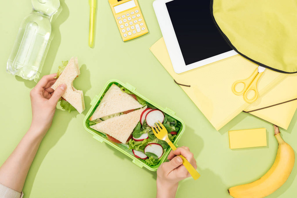 cropped view of woman hands with sandwich, plastic utensils over lunch box with food near backpack, digital tablet, bottle of water and stationery - Photo, Image