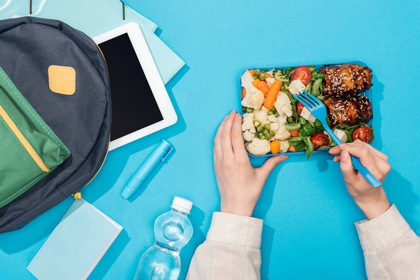 cropped view of woman holding lunch box near backpack with folders, stationery, bottle of water and digital tablet - Photo, Image