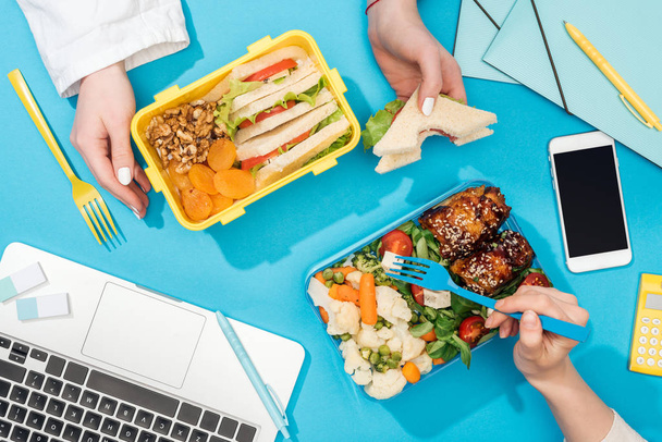 cropped view of two women holding forks over lunch boxes with food near laptop - Photo, Image