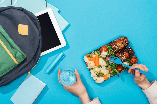 cropped view of woman holding lunch box and glass of water near backpack with folders, stationery and digital tablet - Photo, Image