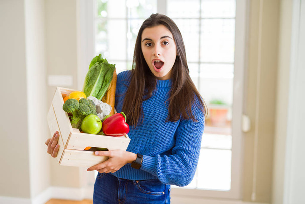 Beautiful young woman holding wooden box full of healthy groceries scared in shock with a surprise face, afraid and excited with fear expression - Photo, Image