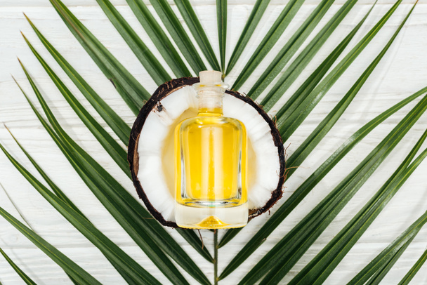 top view of coconut oil in bottle on coconut half and green palm leaf on white wooden surface - Foto, afbeelding